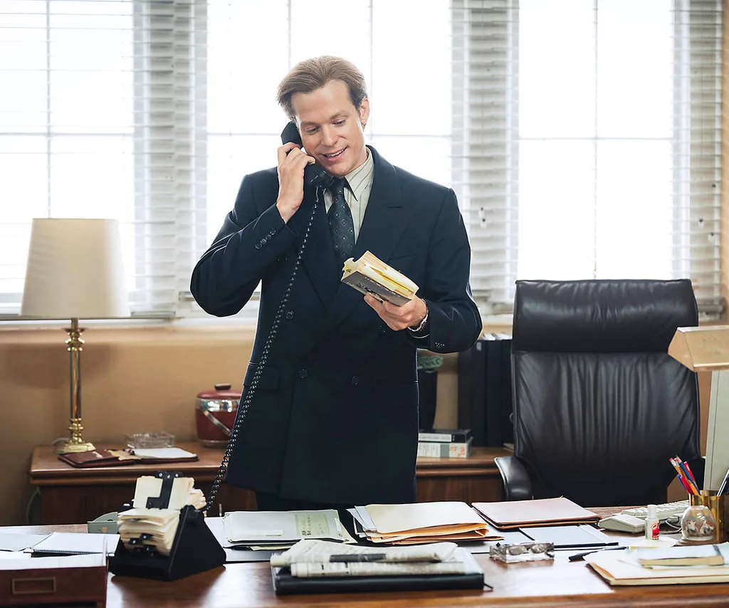 Sam Reid, as Dale Jennings, stands at his desk, holding the phone, in The Newsreader.