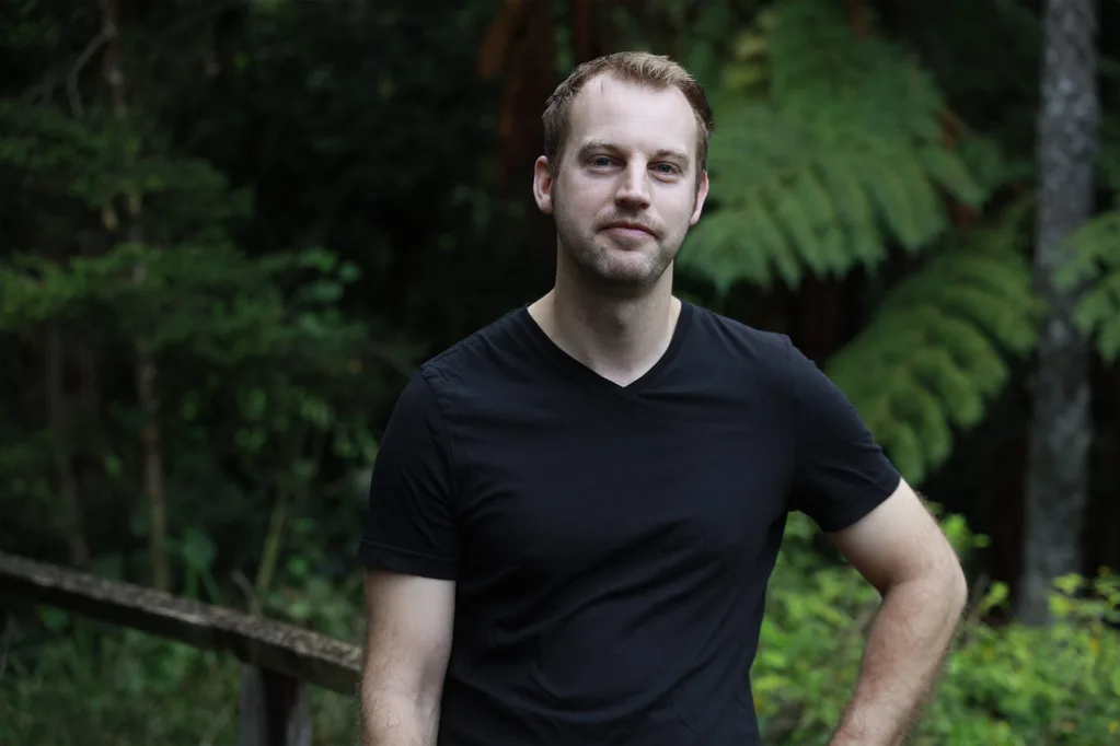 Blond Tim Booth stands in black t-shirt in front of garden