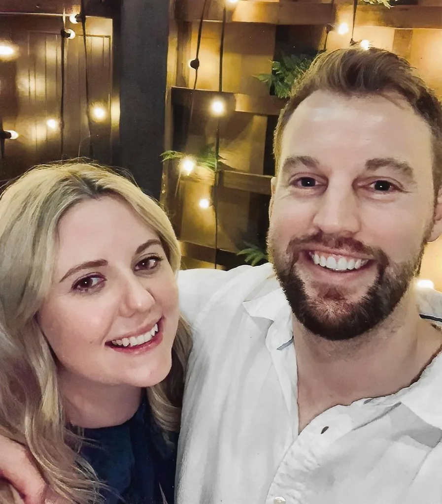 Bearded Tim stands in white shirt with his arm around blonde wifeFiona