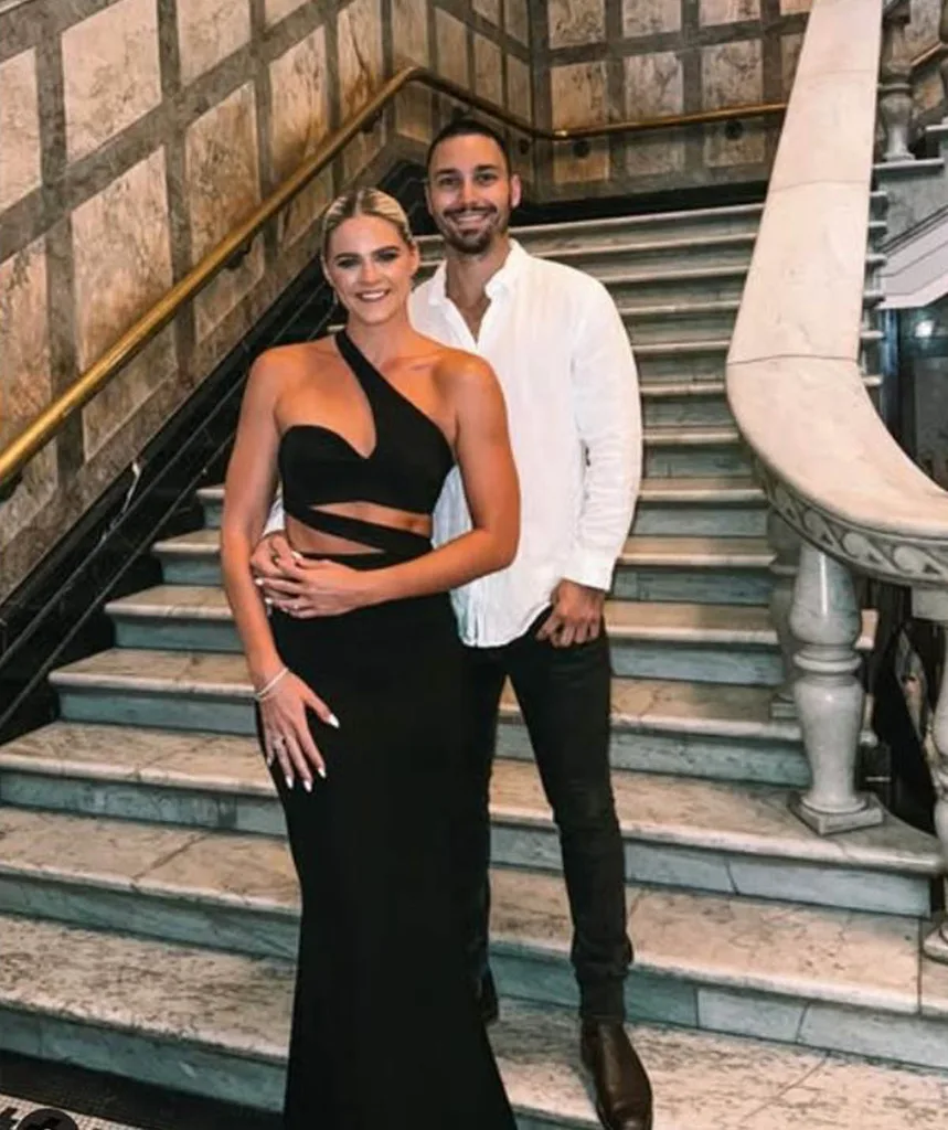 Shayna jack in a stunning black gown partnered with her fiance Joel who is wearing a white button up shirt and black pants, standing at the bottom of a grand staircase.