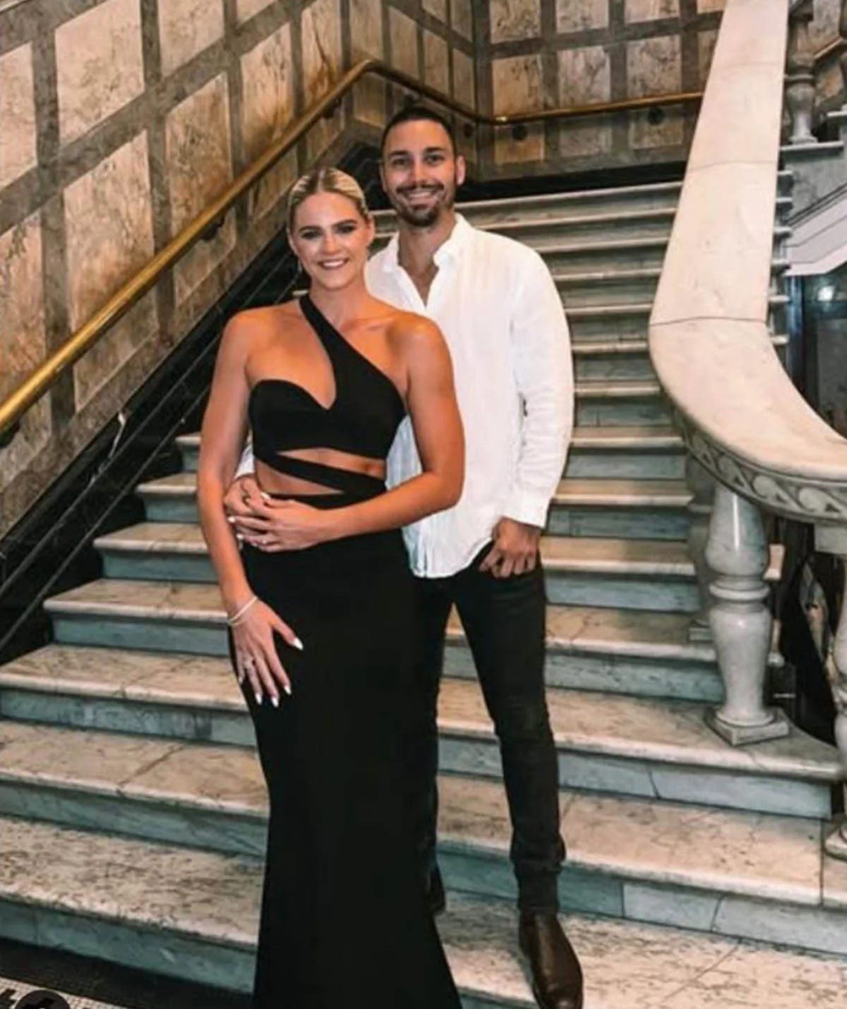 Shayna jack in a stunning black gown partnered with her fiance Joel who is wearing a white button up shirt and black pants, standing at the bottom of a grand staircase.