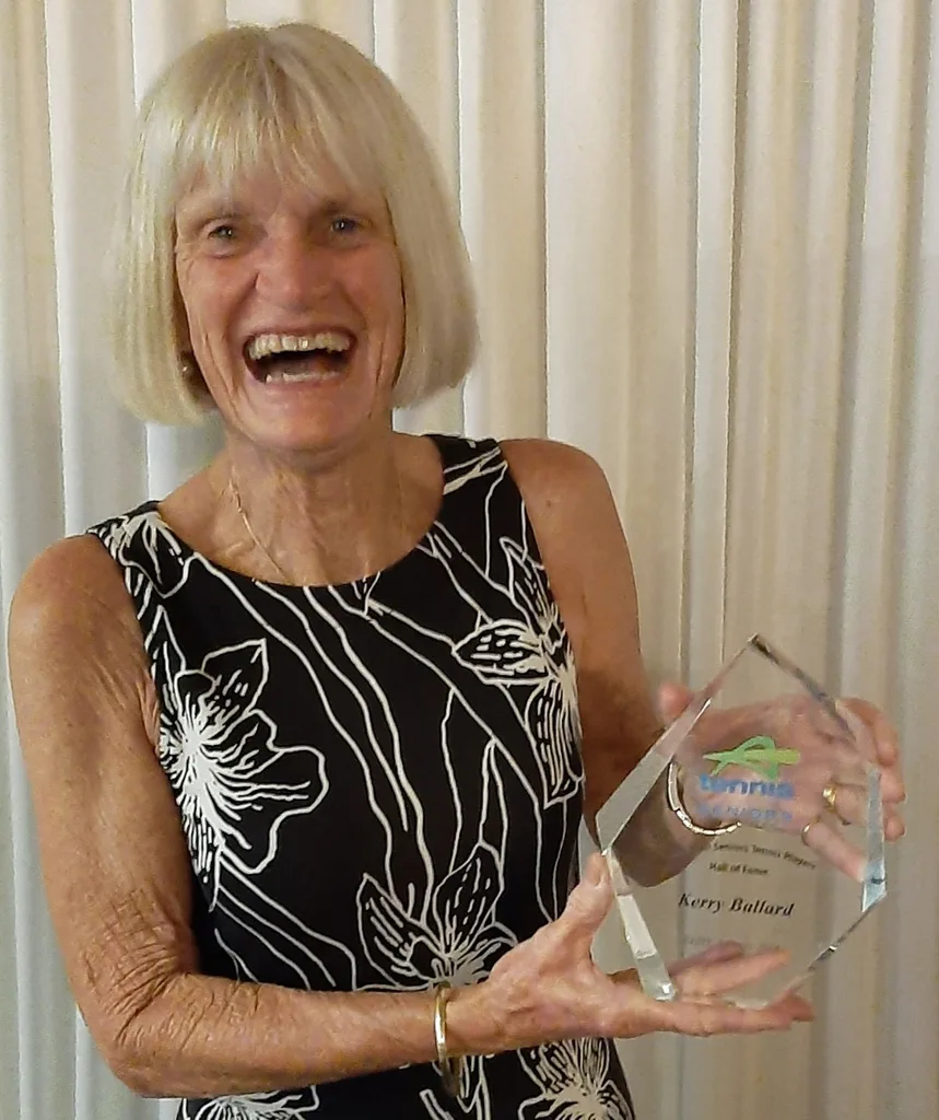 A woman in her seventies with blonde hair is smiling and holding a tennis medal.