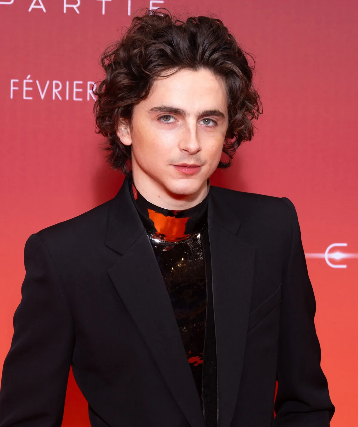 Timothée stands in front of an orange wall in a black suit.