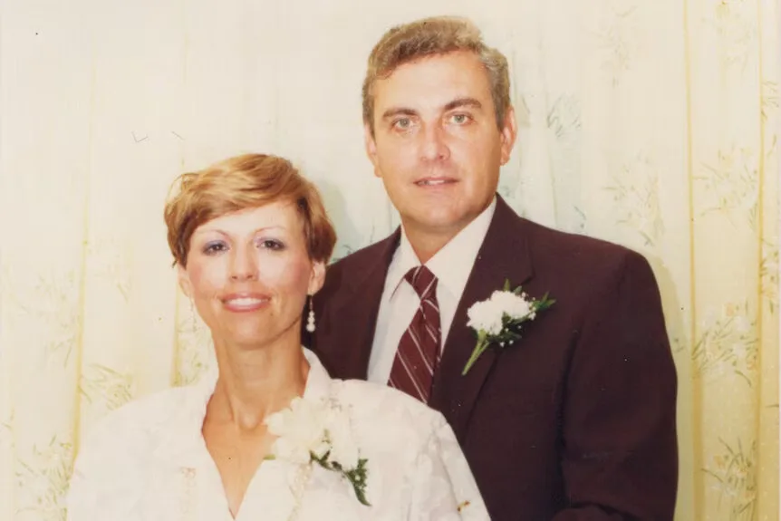 Ken in tux with white flower in his lapel stands behind his wife, with short blonde hair and wearing a white dress