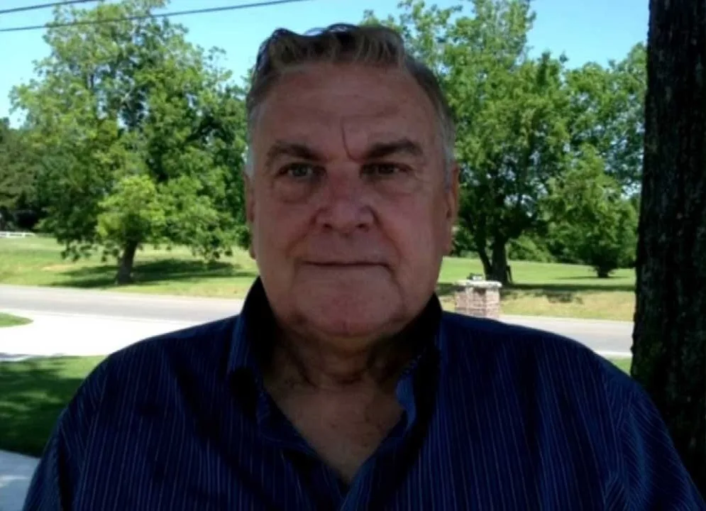 Older Ken in blue coloured shirt in front of green grass and trees