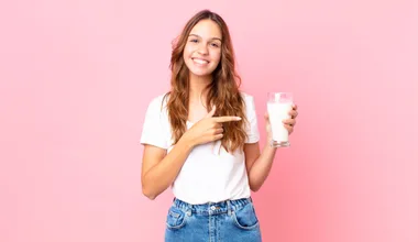 young pretty woman smiling cheerfully, feeling happy and pointing to the side and holding a glass of milk