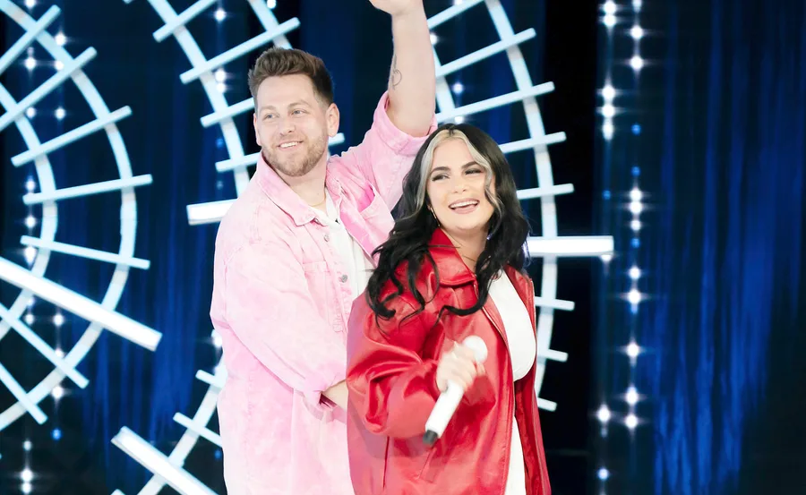 Married couple Chris and Jazz stand next to each other on the Australian Idol audition stage, both smiling.