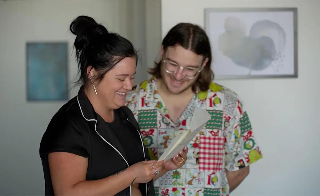Ash in a black shirt with hair a bun smiles as she reads the weekly challenge to her MAFS groom Jake, also smiling with glasses, and in a 'party' shirt.