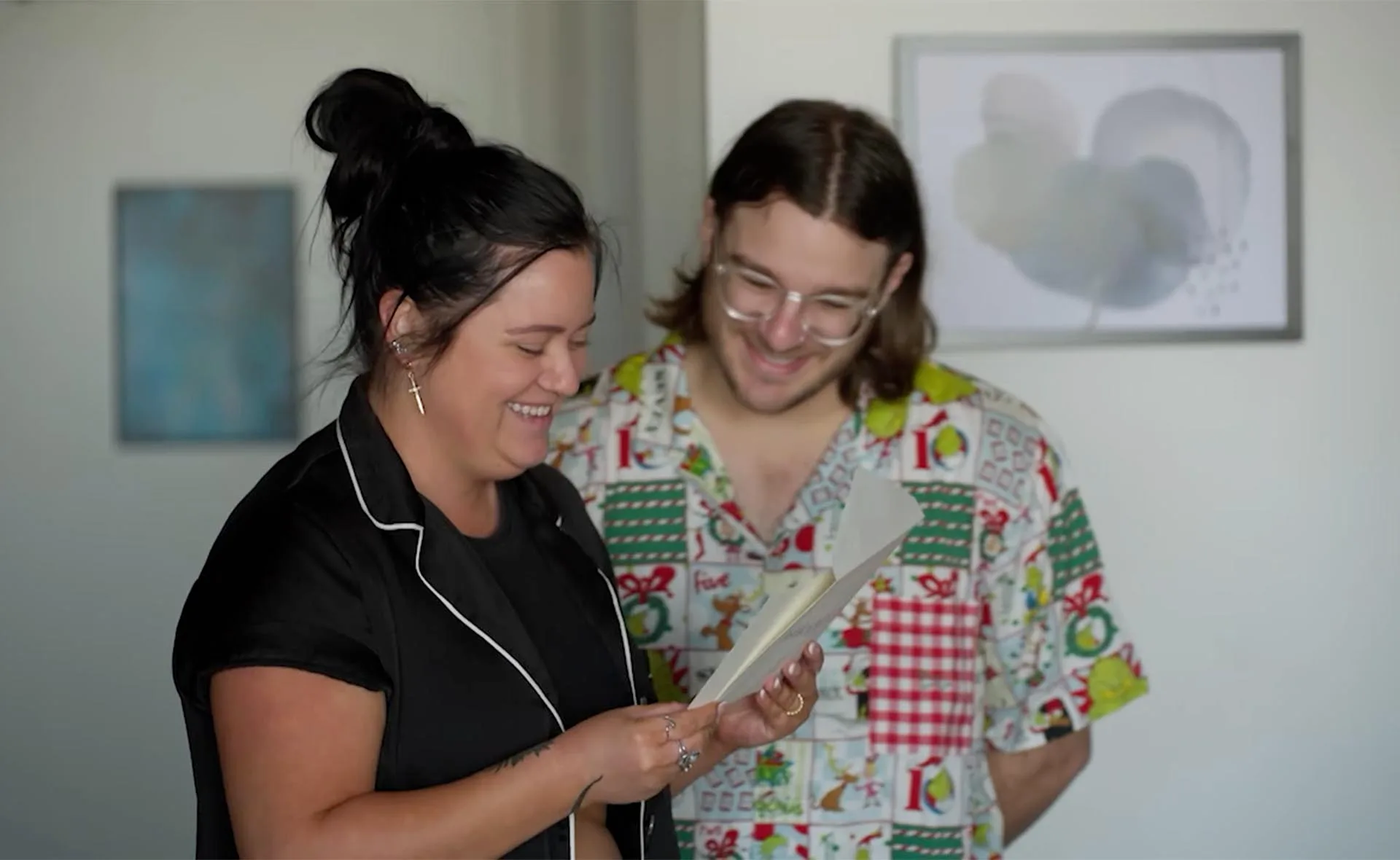 Ash in a black shirt with hair a bun smiles as she reads the weekly challenge to her MAFS groom Jake, also smiling with glasses, and in a 'party' shirt. 