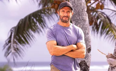 Australian Survivor host Jonathan LaPaglia poses with his arms folded in front of a palm tree