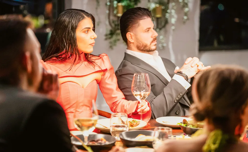Carina in an orange dress at the MAFS dinner table, seated next to her TV husband Paul who is sitting back in his chair. Both have a stern expression on their face.