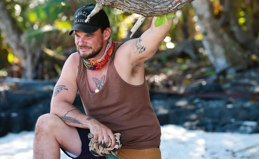 Rich, wearing a singlet, rests up against a tree on the beach in Samoa.