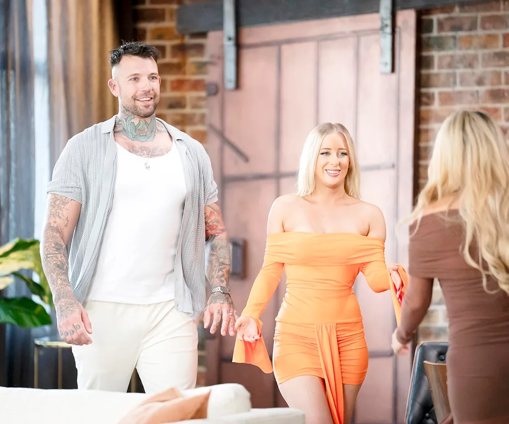 Dave and Jamie enter the dinner party all smiles. Dave in a white shirt, grey over shirt and beige pants. Jamie in a stunning off the shoulder orange dress.