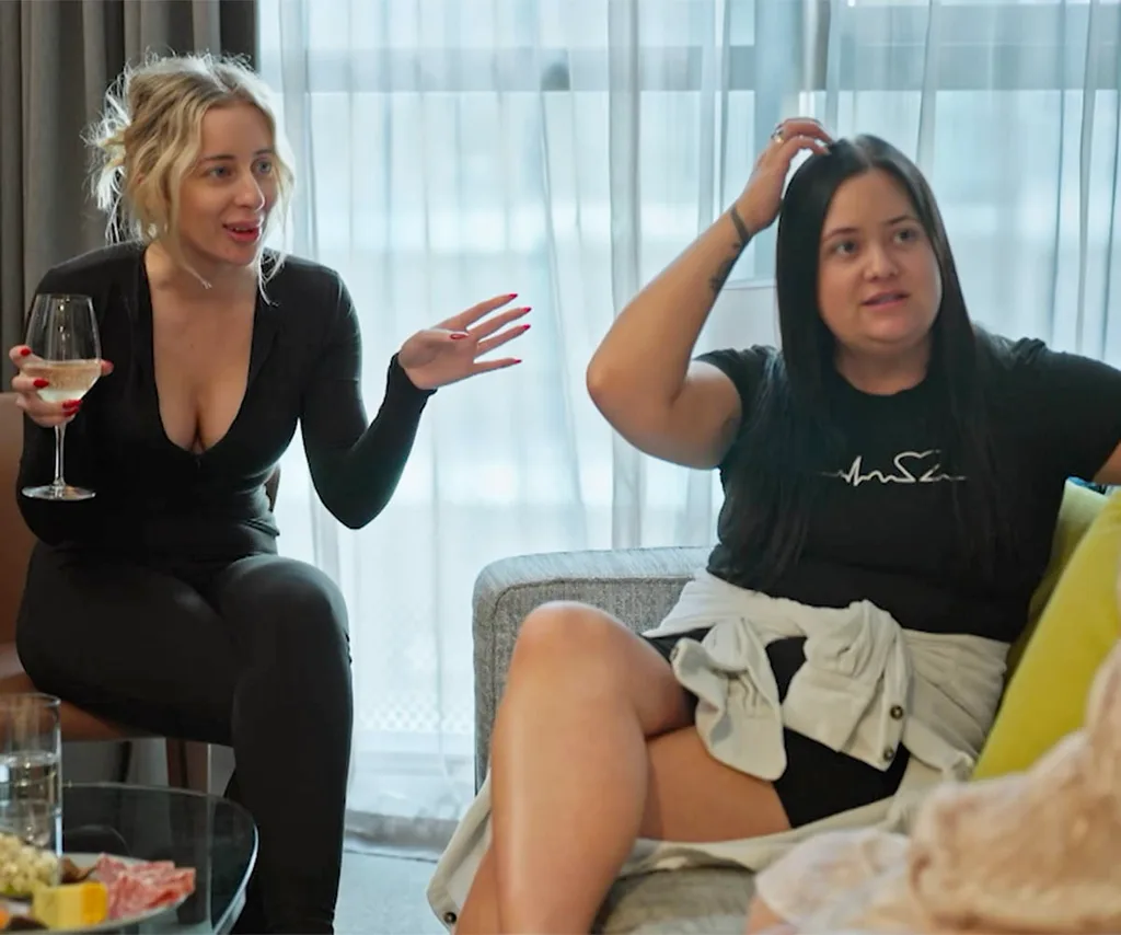 Jamie (left) sits on the couch witha wine glass in hand, Ash is in the middle on a couch talking to Katie as they all debrief about about the disastrous Photo Ranking challenge.