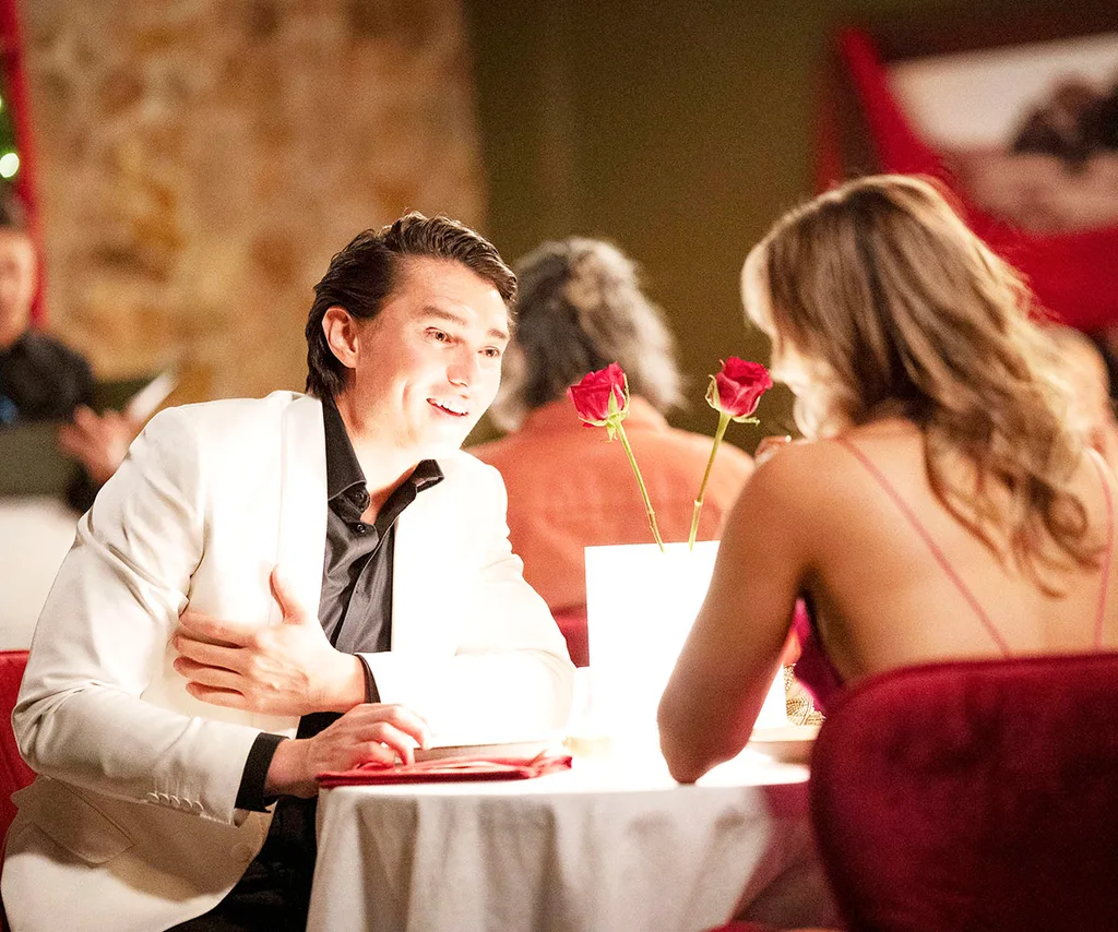 Leo is pictured in a white suit with a black button up looking happy at a table with a white tablecloth, roses, facing Krista. Only the back of her is pictured in a red dress as they attend a romantic dinner.