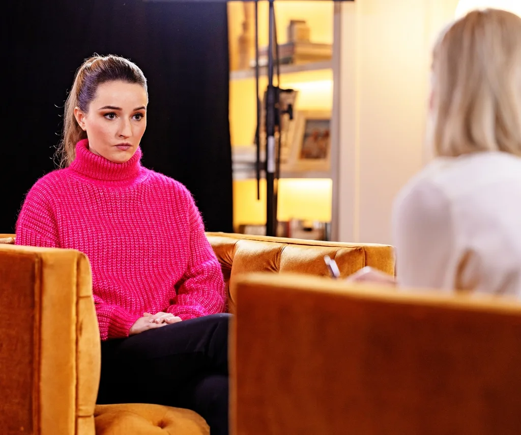 A woman in a bright pink jumper sits opposite a woman interviewing her.