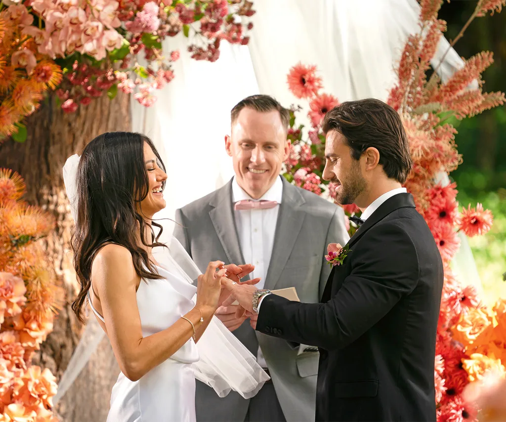 Veronica in a white wedding dress holding hands with Eliot in a suit at the alter with beautiful flowers in the background with a male celebrant in a gray suit