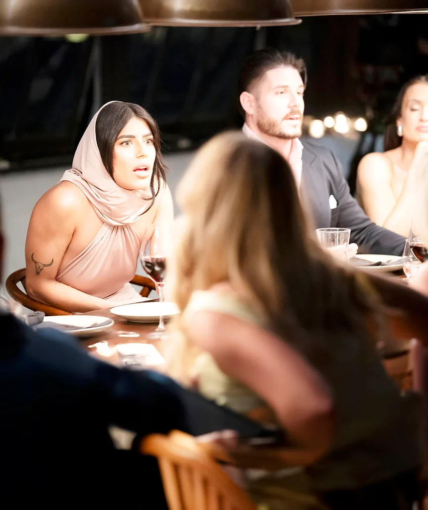 Carina sits leaning across the table with a look of rage and horror across her face. She is seated next to her husband Paul by wearing a beige head scarf fashion statement piece.