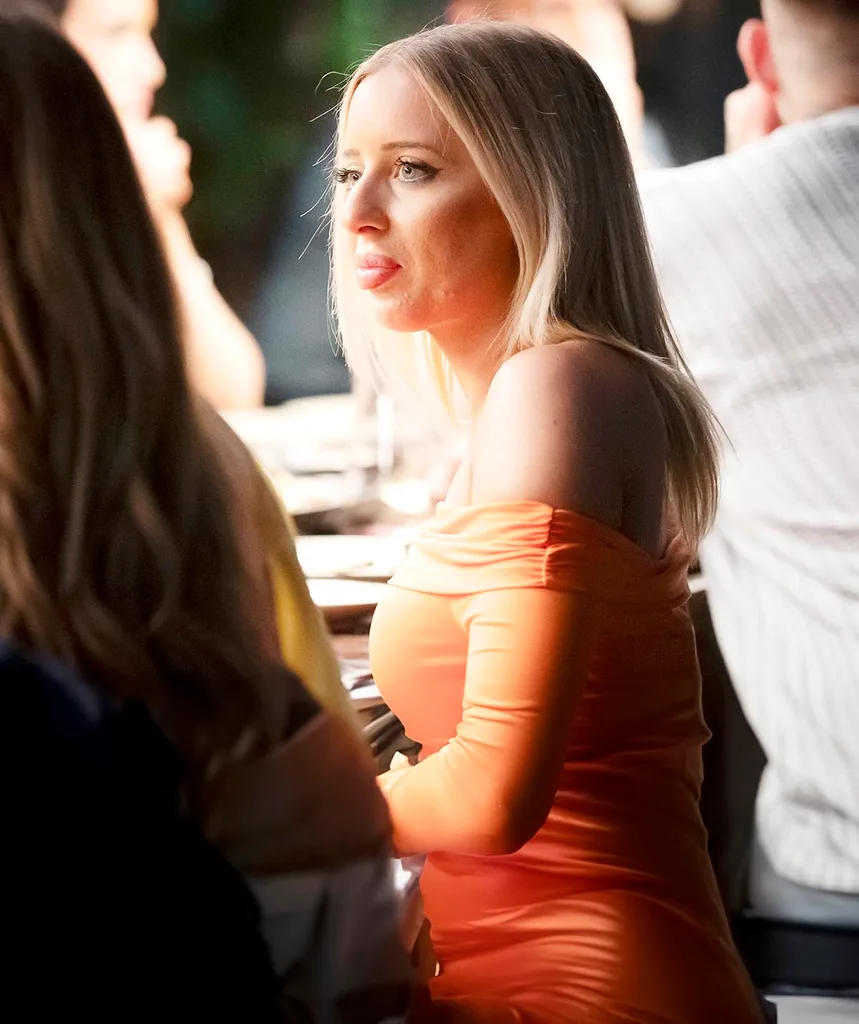 Jamie sits at the dinner party table in an orange dress looking confronted.