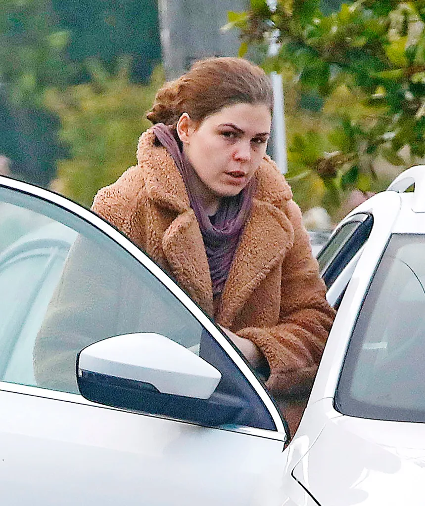 An image of a woman in a brown coat looking downcast, getting into a white car.