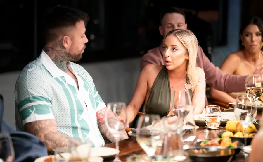 Jamie, wearing an olive green dress, looks intently into the eyes of Dave, who is wearing a patterned shirt, as they sit at the table at the dinner party