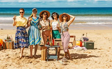 Five women, dressed in 1950s outfits, and wearing hats and sunglasses, stand on the beach and smile for the camera.