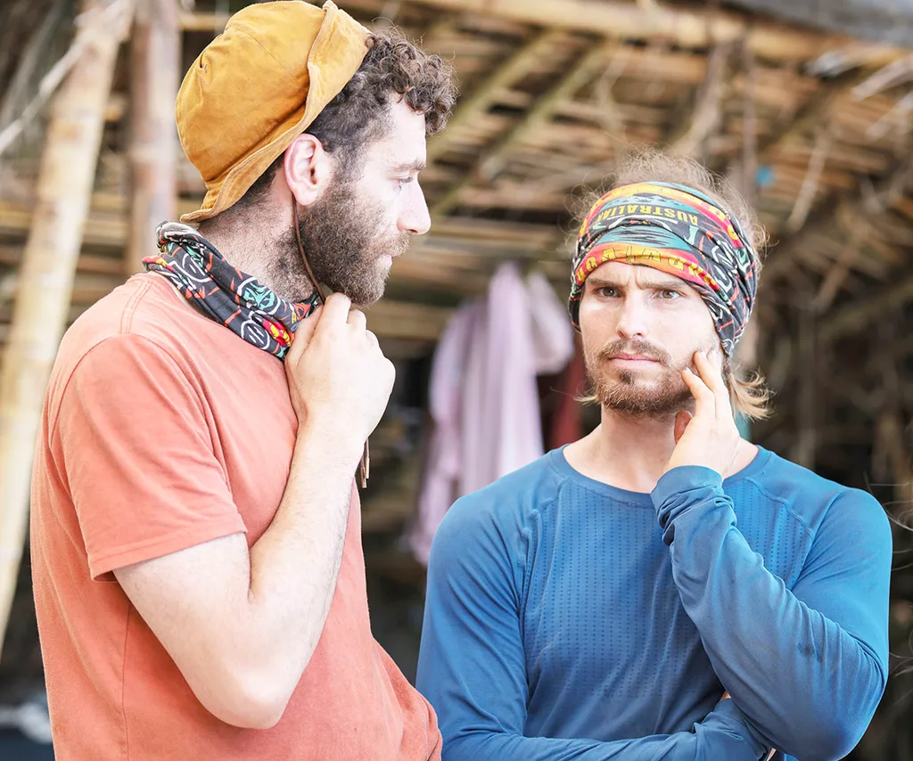 Kaelan is standing next to AJ, looking thoughtful, in front of their shelter in Samoa.