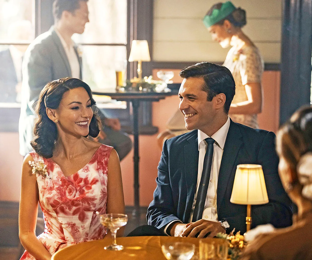 Michelle Keegan, as Kate, wearing a white dress with red flowers, smiles at Nic English, as Robbie, in a restaurant