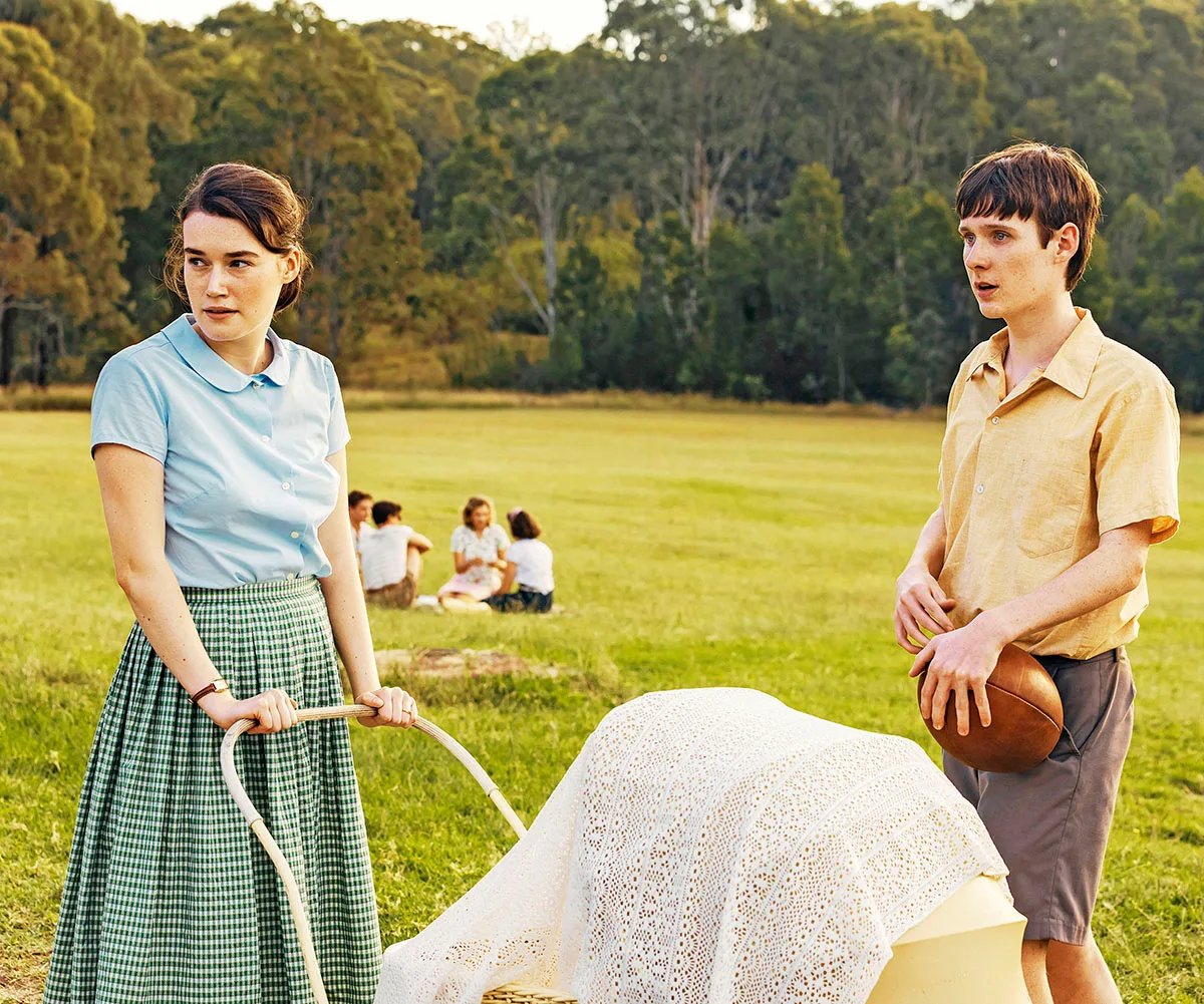 Hattie Hook, as Pattie, wearing a pale blue top and a long skirt, pushes a pram in a park. 