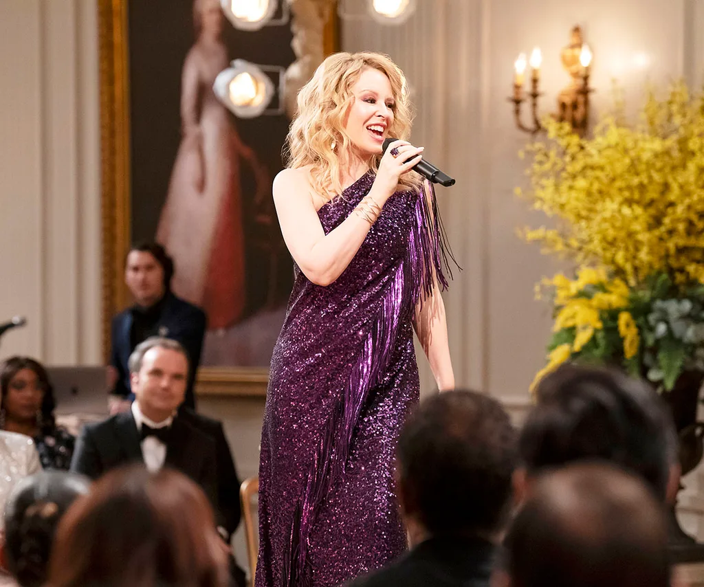 Kylie Minogue, in a purple sparkly dress, sings into a microphone.