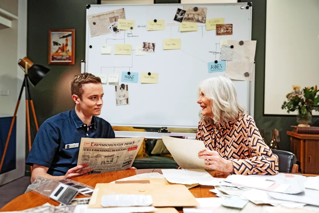 JJ and Agnes sit at a table pouring over historical documents with a white board in the background.
