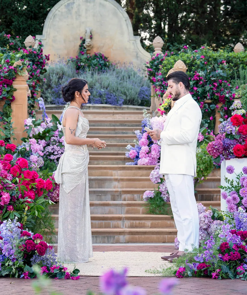 Carina on the left in a stunning gown and Paul on the right in an all white suit. Things look tense as he reads from his Final Vows cards. Their is a staircase in the background and a beautiful display of lavender and flowers in the foreground.