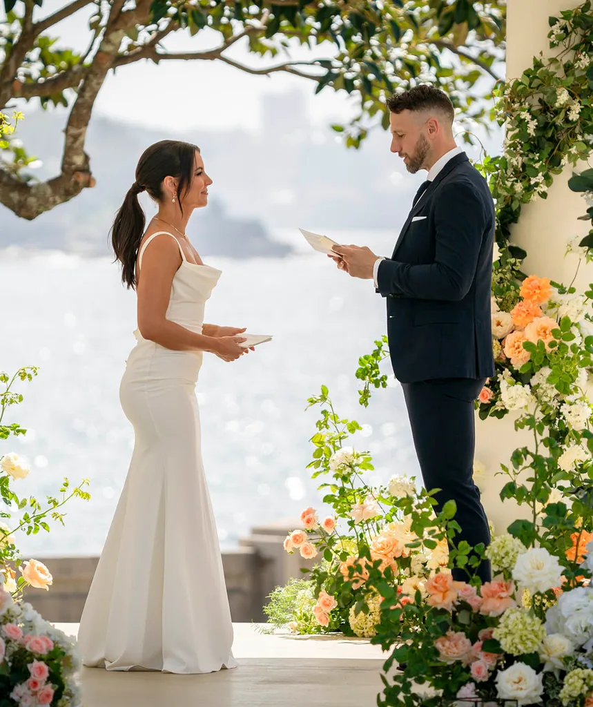 Rhi on the left in a white dress and Jeff on the right suited up as he reads from his Final Vows cards. Their is the sea in the background and flowers in the foreground.