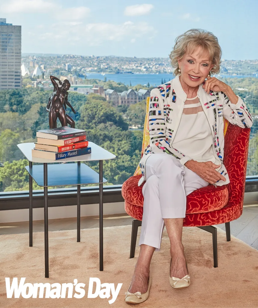 Blanche sits on an orange chair in her apartment with the view of the Sydney harbour in the background.