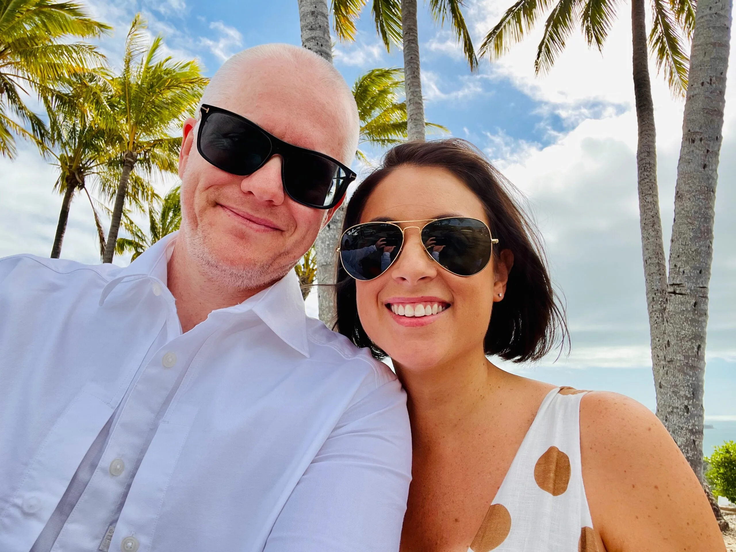 Daniel and Jade wearing sunglasses standing beneath palm trees and blue skies