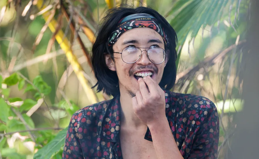 Myles, wearing his buff on his head, looks upwards on the beach in Samoa, with a smile on his face