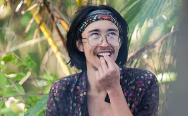 Myles, wearing his buff on his head, looks upwards on the beach in Samoa, with a smile on his face