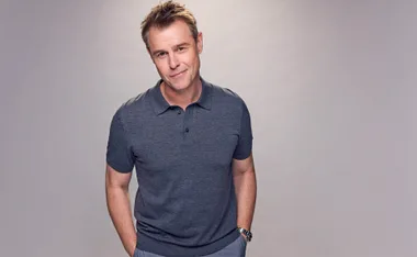 Rodger Corser pictured in a grey polo with his hands in his pockets against a light grey backdrop.