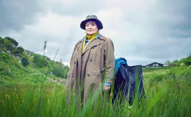 Vera in her famous trench coat with a hat in green rolling hills with weed in the foreground