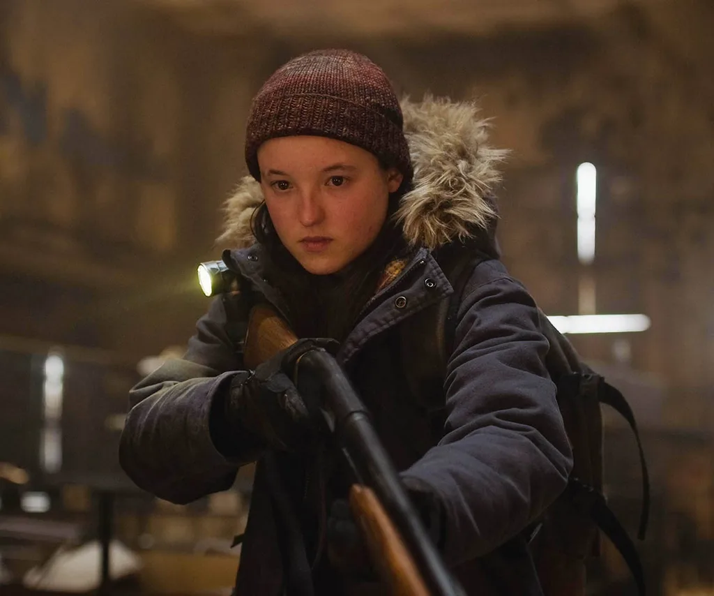 Ellike points a shotgun while wearing a snow jacket and beanie in a barn