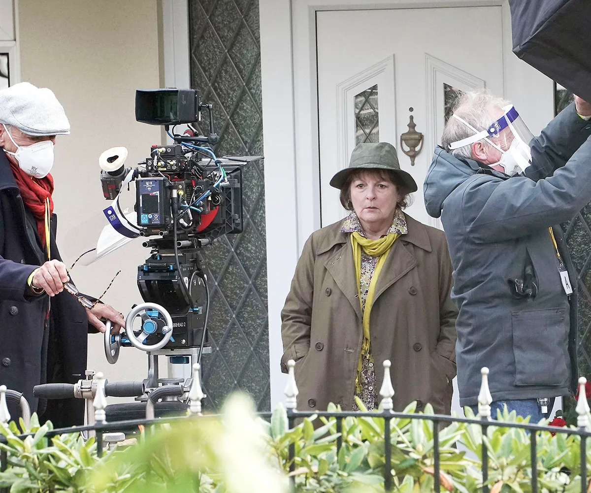Vera is shot in front of a white door to a home while cameramen with Covid masks on film around her