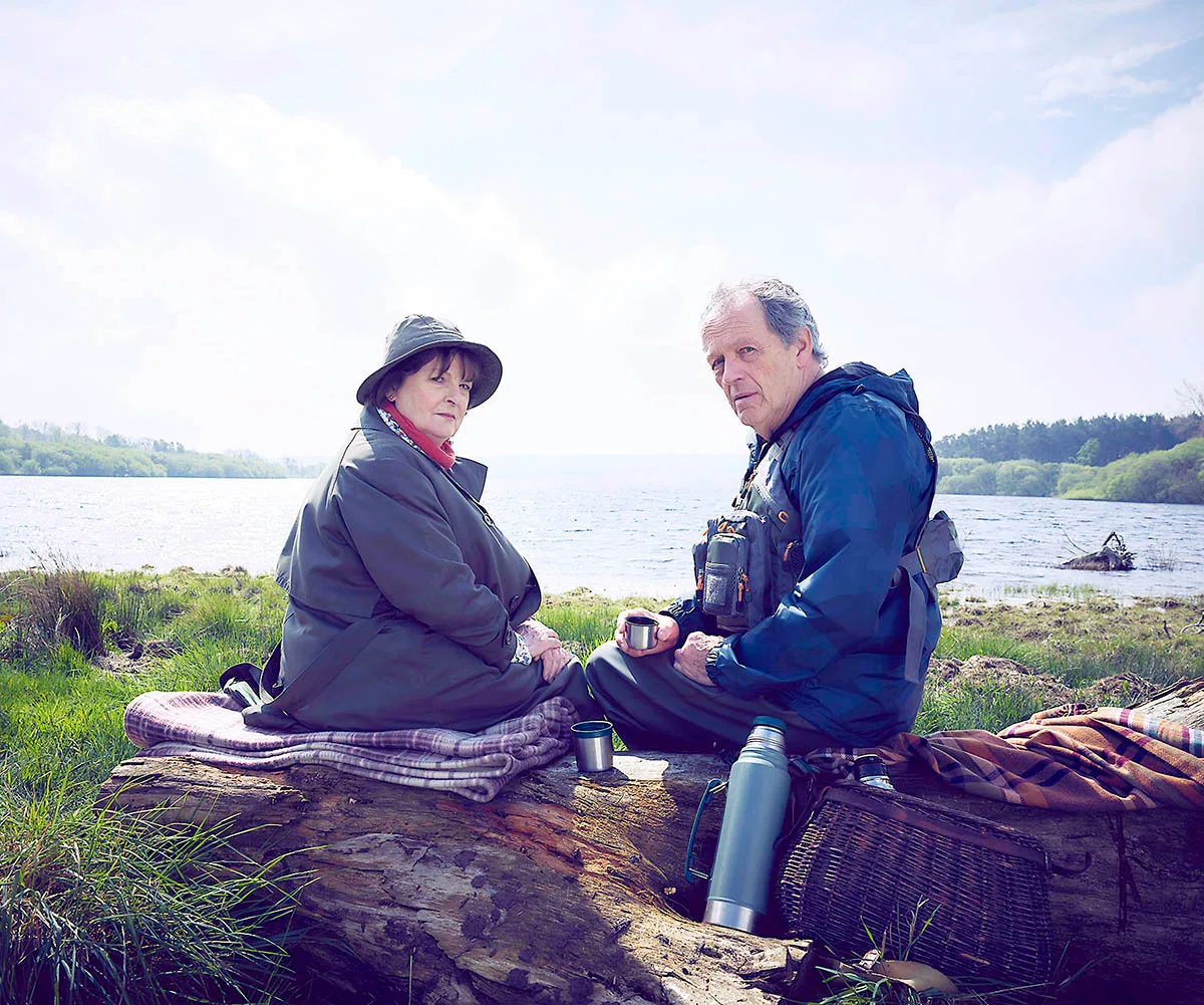 Brenda Blethyn and Kevin Whately sit on a log and have a picnic on the grass with a lake in the background
