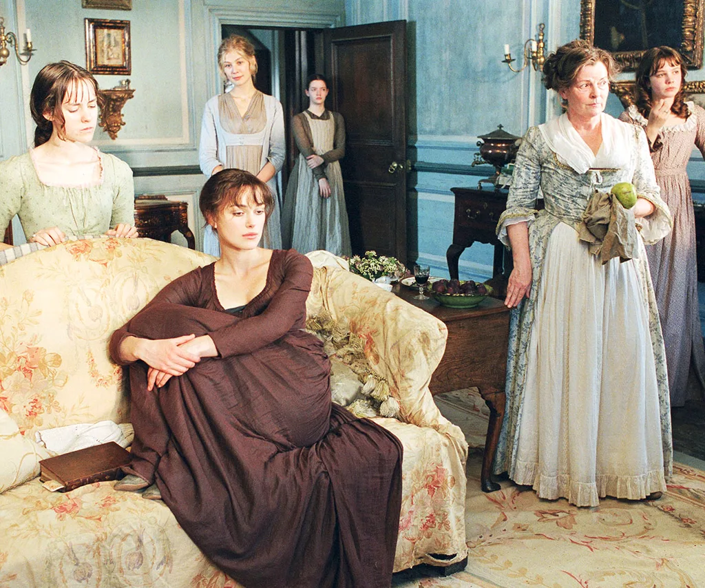 An image from the film Pride and Prejudice of a room full of ladies in a rich Victorian living room while some stand in their old style dresses and another sits on the couch