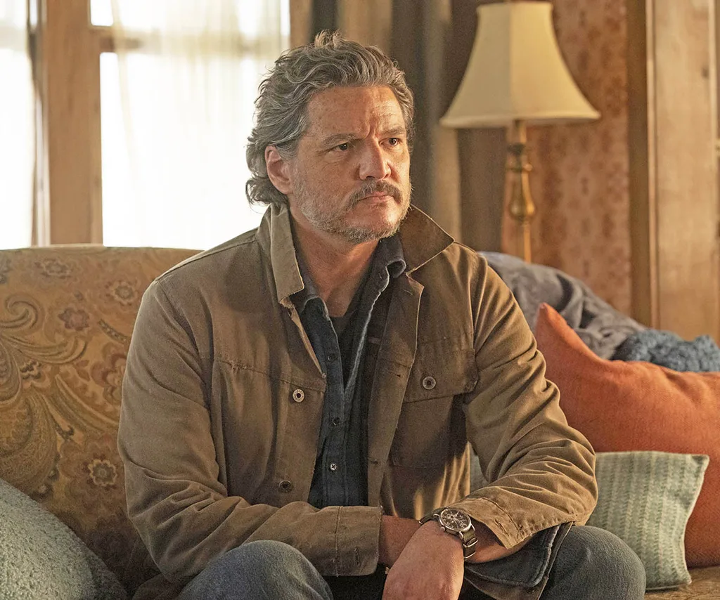 Pedro Pascal sits on a couch in a brown jacket and looks somber