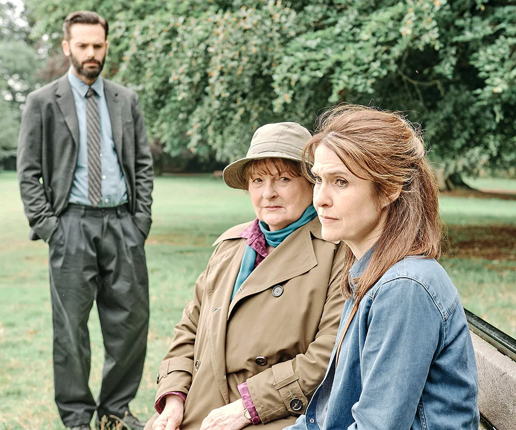 Joe Ashworth stands in a suit looking glum while Vera and a woman sit on a bench.