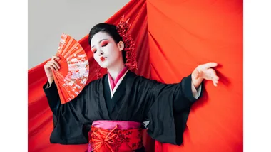 A Japanese geisha poses artfully with a fan dressed in a black kimono.