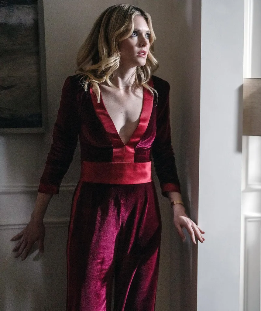 Meghann is backed against a wall hiding from a killer looking distressed in a red velvet low cut long sleeve jumpsuit