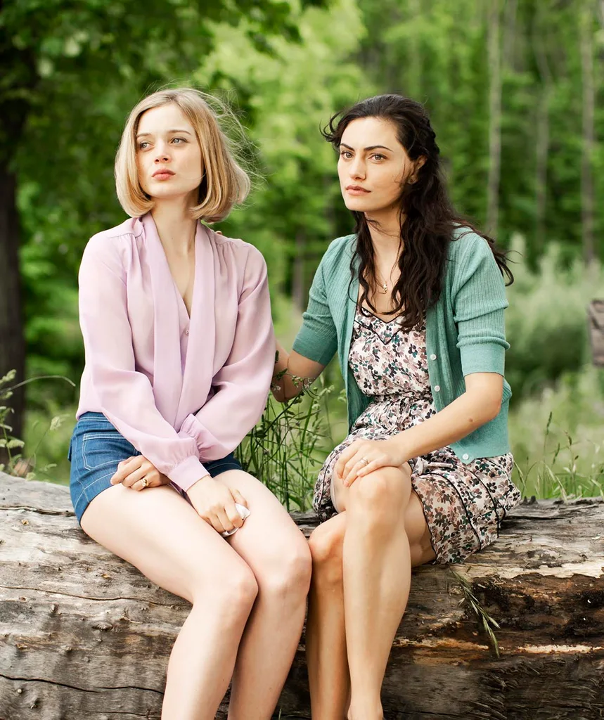 Bella and Phoebe sit on a log in the wilderness and Phoebe has a hand on Bella's back comforting her as they both look off into the distance