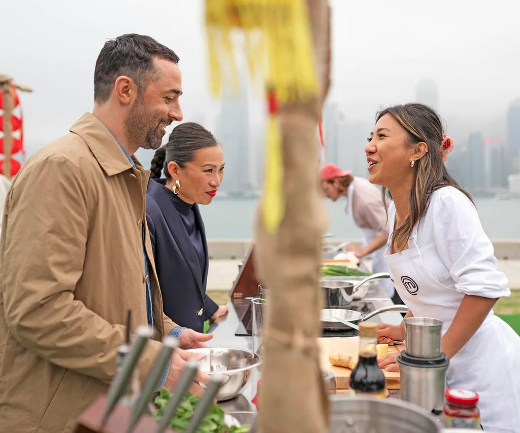 Andy and Poh smile as they give advice to a grinning contestant at a challenge with the banks of Melbourne city and the Yarra river in the background