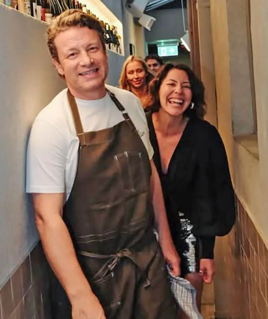 Jamie Oliver in a white tee and an apron is pictured in a hallway with Sofia Levin smiling